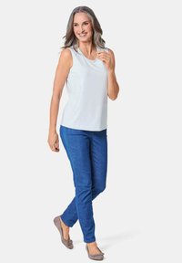 Top bianco senza maniche con scollatura rotonda, abbinato a jeans blu a taglio slim e scarpe ballerine grigie. Il modello è in una posa informale.