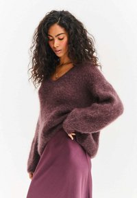 Bordowy, puszysty sweter z dekoltem w serek, długimi rękawami i luźnym krojem, zestawiony z gładką, fioletową spódnicą. Miękka tekstura i swobodny design.