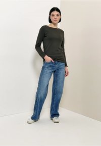 Olivengroen shirt met lange mouwen gecombineerd met lichtblauwe jeans met rechte pijpen. Model staat voor een neutrale achtergrond en draagt witte sneakers.