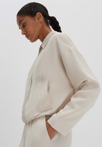 Helle beiges Bomberjacke mit Frontreißverschluss, Seiten Tasche, lockerer Passform und glattem Stoff. Verfügt über einen Stehkragen und elastische Bündchen.
