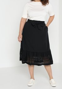 Svart midikjol med volang i nederkant, med intrikata spetsdetaljer. Stiligt kombinerad med en vit t-shirt med kort ärm och avslappnade vita sneakers.