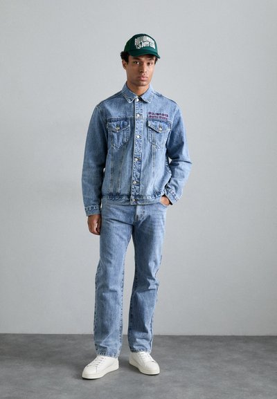 Veste en denim bleu clair avec deux poches poitrine et un logo. Associée à un jean bleu clair et des baskets blanches, surmontée d'une casquette verte.