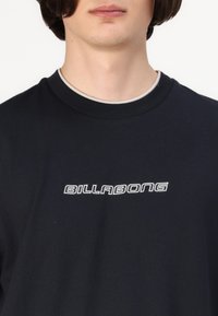 Juodas medvilnės marškinėliai su apvali apykakle, ant krūtinės esantis baltas "BILLABONG" logotipas išraiškingu šriftu.