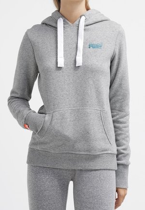 Personne portant un sweat à capuche gris clair avec des cordons blancs et un pantalon gris assorti, une main dans la poche avant.