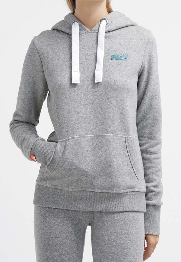 Personne portant un sweat à capuche gris clair avec des cordons blancs et un pantalon gris assorti, une main dans la poche avant.