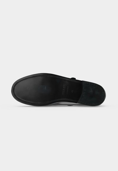 Semelle de chaussure en cuir noir avec bords cousus et détails cloutés au talon, présentée sur un fond clair uni.