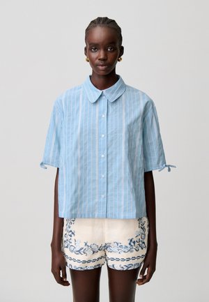 Femme portant une chemise boutonnée à rayures verticales bleu et blanc et un short blanc avec une broderie florale bleue, debout devant un fond uni.