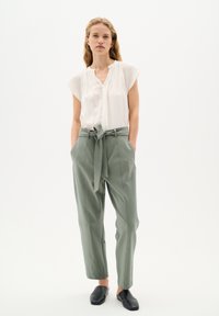 Blusa bianca a maniche corte con pieghe, abbinata a pantaloni alto vita di un verde chiaro con cintura annodata e tasche laterali. Scarpe nere senza lacci.