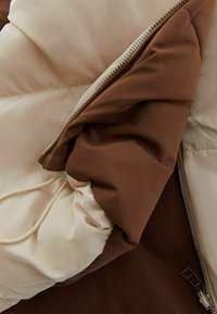 Giacca a vento con una combinazione di colori beige e marrone, dotata di chiusura con zip, dettagli con cordoncino e una morbida texture trapuntata.