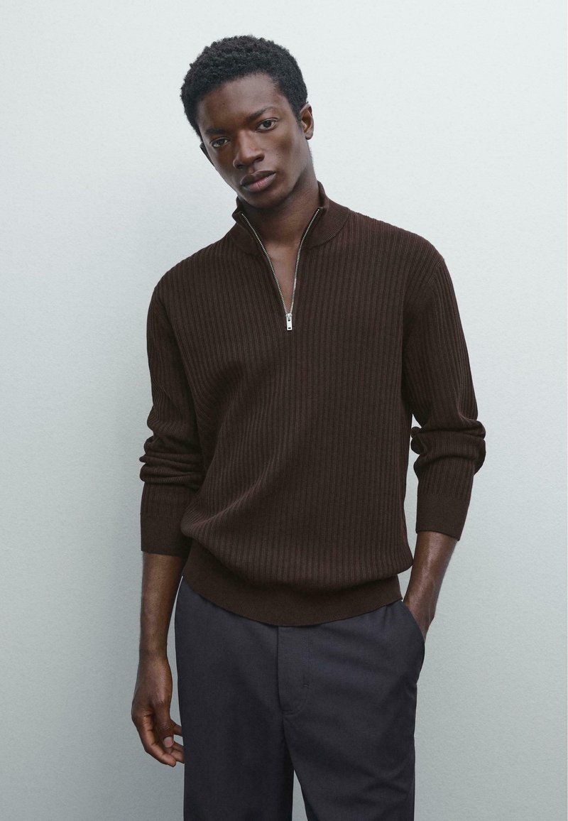 Brauner gerippter Pullover mit halbem Reißverschluss und enganliegenden Manschetten, kombiniert mit dunklen Hosen. Weiche Textur, lässiges Design.