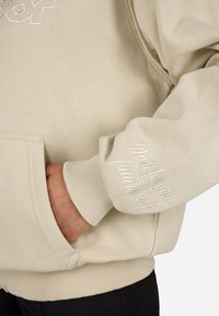 Beige kapucnis pulóver hímzett szöveggel az ujjon, kenguruzsebbel, bordás mandzsettákkal és puha, texturált anyagból. Egyszerű és praktikus kialakítás.