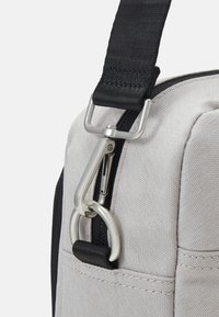 Ecke einer grauen Stofftasche mit schwarzem Tragegurt, ausgestattet mit einem silbernen Metallhaken und einem Reißverschluss; glatte Textur und sichtbare Steppnähte.