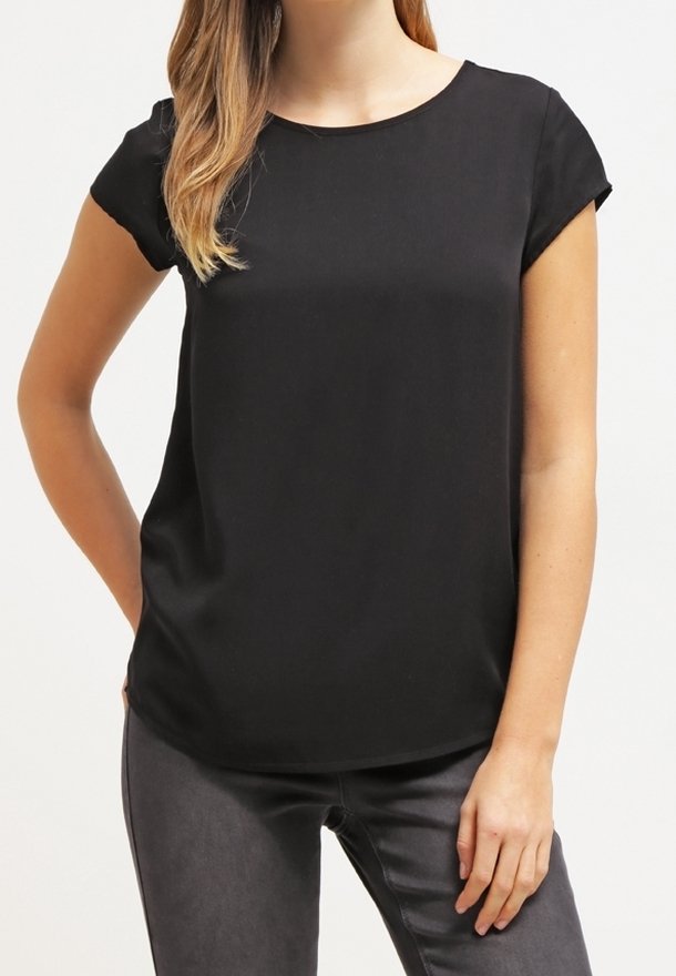 Femme portant un chemisier noir uni à manches courtes et un pantalon gris foncé, debout avec une main sur la hanche et l'autre détendue.