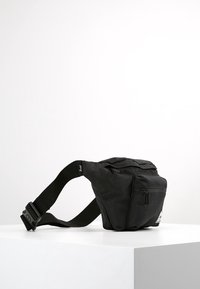 Černá látková ledvinka s nastavitelným popruhem, zipovými kapsami a plochým profilem. Obsahuje logovou nášivku a hladkou texturu.