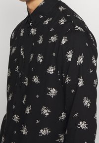 Camicetta nera con motivo floreale bianco, dotata di colletto, bottoni davanti e una vestibilità comoda. Realizzata in tessuto leggero.