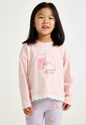 Nuori tyttö, jolla on suorat mustat hiukset, pukeutuneena vaaleanpunaiseen pitkähihamaiseen toppiin, jossa on söpö juoma- ja leivonnaiskuvio, yhdistettynä pastellivärisiin kuvioituihin legginsseihin.