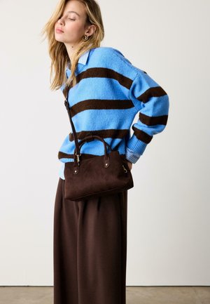 Giovane donna che indossa un maglione a righe blu e marrone, pantaloni marroni a gamba larga e porta una borsa a tracolla marrone scuro.