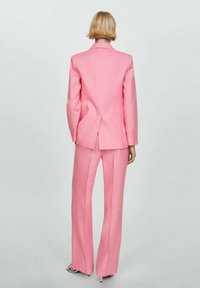 Svetlo roza prilagojeni kostim, ki vključuje oprijet blazer z enim odprtjem in širokimi hlačami. Izdelan iz gladkega materiala, z minimalnimi detajli.