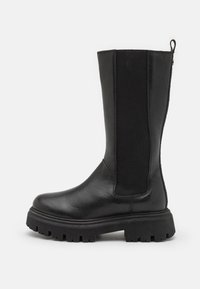 Botte noire hauteur genou avec un extérieur en cuir lisse et des panneaux élastiques sur les côtés. Elle dispose d'une semelle en caoutchouc épaisse avec des profondes rainures pour une adhérence optimale.