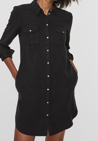 Black button-up shirt dress with a collar, two chest pockets, rolled sleeves, and a curved hem. Made of a smooth fabric.