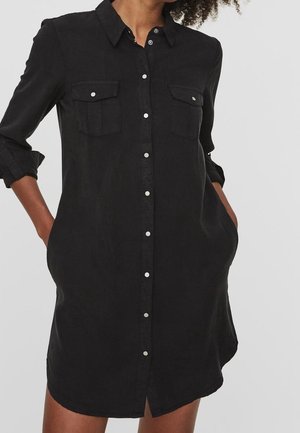 Black button-up shirt dress with a collar, two chest pockets, rolled sleeves, and a curved hem. Made of a smooth fabric.