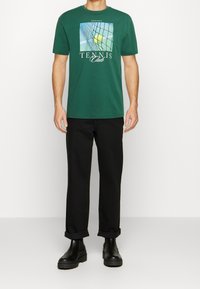 Grön bomulls-T-shirt med tryck av ett tennisnät och logotexten "TENNIS Club" i vitt. Kombineras med uppvikta svarta byxor och kraftiga svarta st övlar.