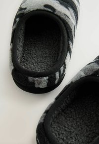 Grau und schwarz gemusterte Hausschuhe mit einer weichen Textur, abgerundeten Öffnungen und einem dunklen Fleece-Futter. Die schwarzen Gummisohlen stehen im Kontrast zum Obermaterial.