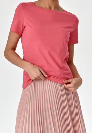 Kvinde iført en koralfarvet top med korte ærmer og et lyserødt, højtaljet plisseret skørt, der holder i toppen kant.