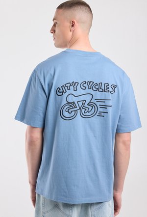 Man met kort haar die een lichtblauw T-shirt draagt met een zwarte "CITY CYCLES"-afbeelding waarop een fietser op de achterkant te zien is.