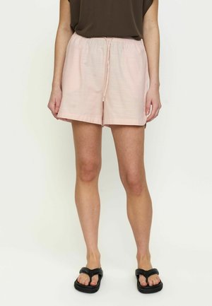 Žena oblečená v svetloružových šortkách so sťahovacou šnúrkou, čiernych žabkách a tmavom bezrukávovom topu, stojaca na svetlom pozadí.