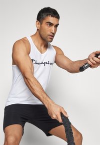 Camiseta sin mangas deportiva blanca con el logo de "Champion", combinada con pantalones cortos negros. La persona sostiene una soga de batalla negra, mostrando unos brazos musculosos.