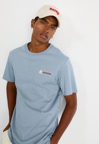 Lyseblå kortærmet t-shirt med et Converse logo-patch og en rund halsudskæring, parret med en beige kasket med rød tekst.