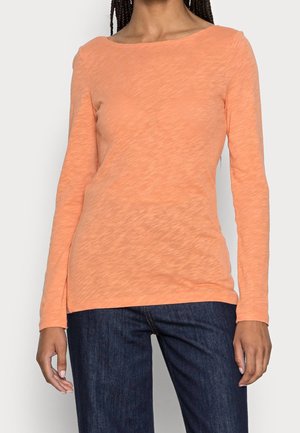 Femme portant une chemise texturée à manches longues de couleur orange clair et un jean bleu foncé, debout avec les bras détendus sur les côtés.