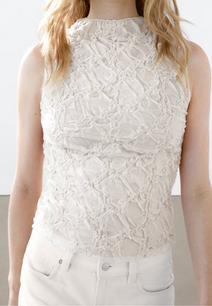 Kobieta w białym, bezrękawnym topie z wysokim kołnierzem i marszczoną kwiatową fakturą oraz białych spodniach, pokazana od ramion do bioder.