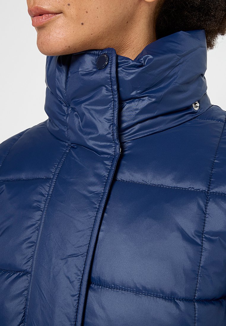 Giacca a vento blu navy con colletto alto, caratterizzata da una texture liscia e lucida e da bottoni a scatto sul colletto. Motivo a quadretti cucito.