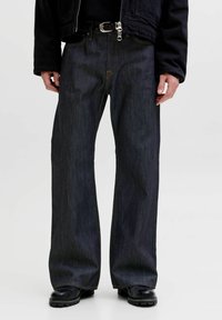 Person iført mørkeblå vidde jeans, sort bælte, sort jakke og sorte støvler, stående mod en ensfarvet lys baggrund.