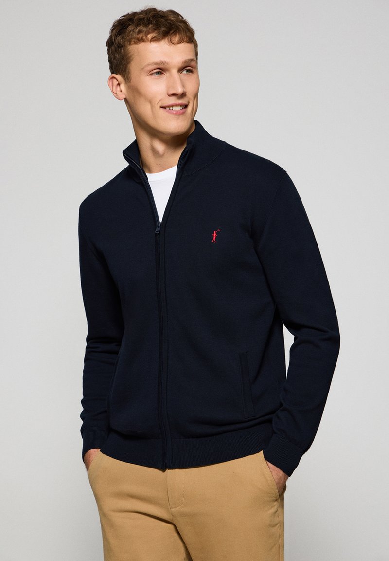 Marinblå zip-up tröja gjord av mjukt material, med hög krage, sidofickor och en liten röd logotyp på bröstet.