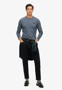 Langsleeve blauwe shirt, donkere jeans en zwarte schoenen. Groene geruite shirt om de taille geknoopt. Zilveren horloge om de pols.