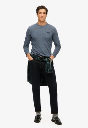 Langsleeve blauwe shirt, donkere jeans en zwarte schoenen. Groene geruite shirt om de taille geknoopt. Zilveren horloge om de pols.
