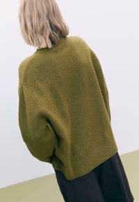 Olive green oversize sweter w miękkim, teksturowanym splocie, z opuszczonymi ramionami i prążkowanym dołem, zestawiony z ciemnymi spodniami.