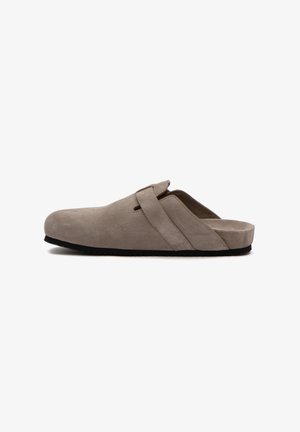 Beige Wildleder-Slipper mit verstellbarem Riemen, geschlossenem Zehenbereich und schwarzer Sohle, seitlich auf weißem Hintergrund gezeigt.