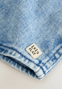 Tissu en denim bleu clair, plié avec une finition texturée. Présente une étiquette en tissu cousue indiquant "JOUONS" en lettres noires.