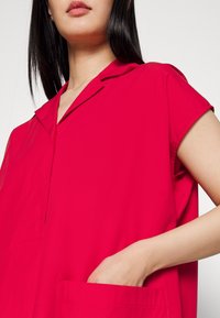 Femme portant une chemise rouge vif à manches courtes avec col, partiellement visible du cou à la taille, main reposant dans la poche avant.