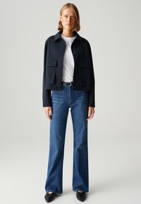 Marineblauwe jas met een kraag en patchzakken, gecombineerd met blauwe rechte jeans en zwarte spitse schoenen.