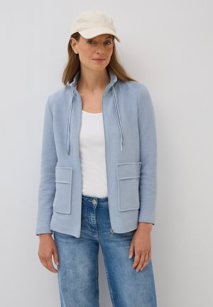 Veste zippée bleu clair avec deux poches avant, tissu texturé et capuche à cordon, portée sur un top blanc et un jean bleu.