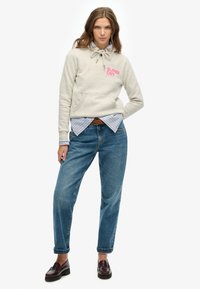 Lichtgrijze hoodie met trekkoorden en "SUPER DRY" logo, gelaagd over een blauw-wit gestreept shirt, gecombineerd met losse blauwe jeans en donkere schoenen.