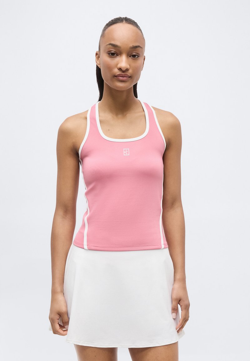 Γυναίκα που φοράει ροζ αμάνικη μπλούζα με λευκά περιθώρια και μικρό λογότυπο, καθώς και λευκή φούστα τένις, πάνω σε απλό ανοιχτόχρωμο φόντο.