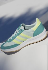 Einzelner türkisfarbener und hellgelber Laufschuh mit weißen Schnürsenkeln und Gummisohle, dargestellt vor einem hellen Hintergrund mit Schatten.