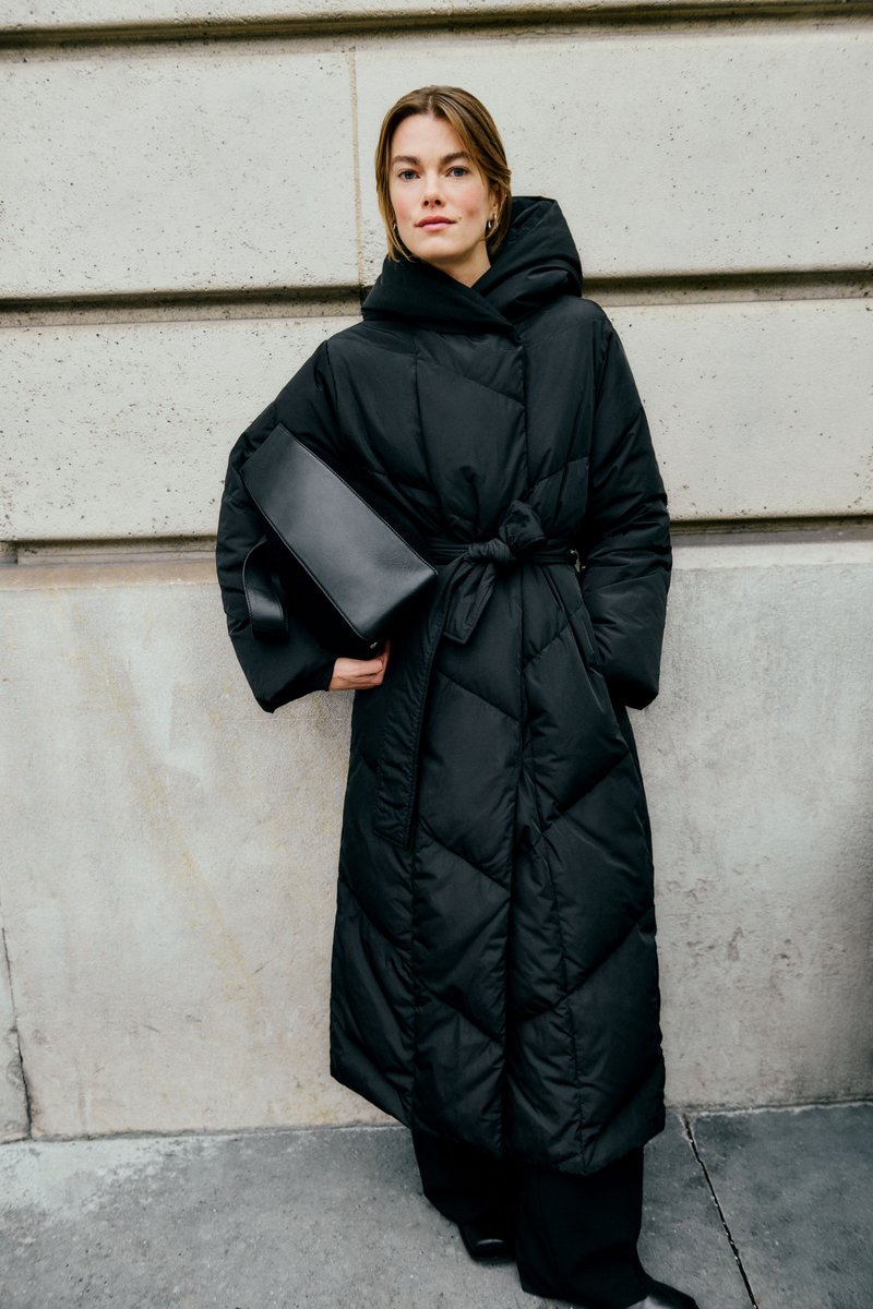 Donna con cappotto lungo e nero imbottito, con vita annodata e cappuccio, che tiene una grande pochette nera, in piedi contro un muro di pietra.