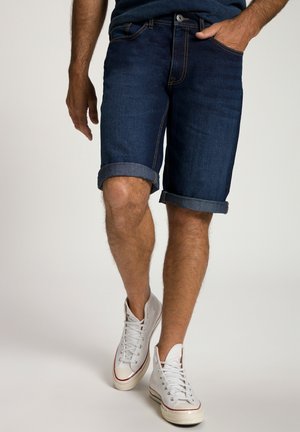 Donkerblauwe denim shorts met opgerolde zomen, voorzien van een klassieke vijf-pocketontwerp en contrasterende stiksels. Gepaard met witte canvas sneakers.